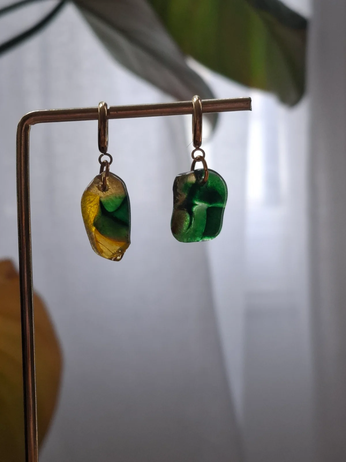 Boucles d'oreilles asymétriques en verre vert et jaune suspendues à un présentoir métallique doré.