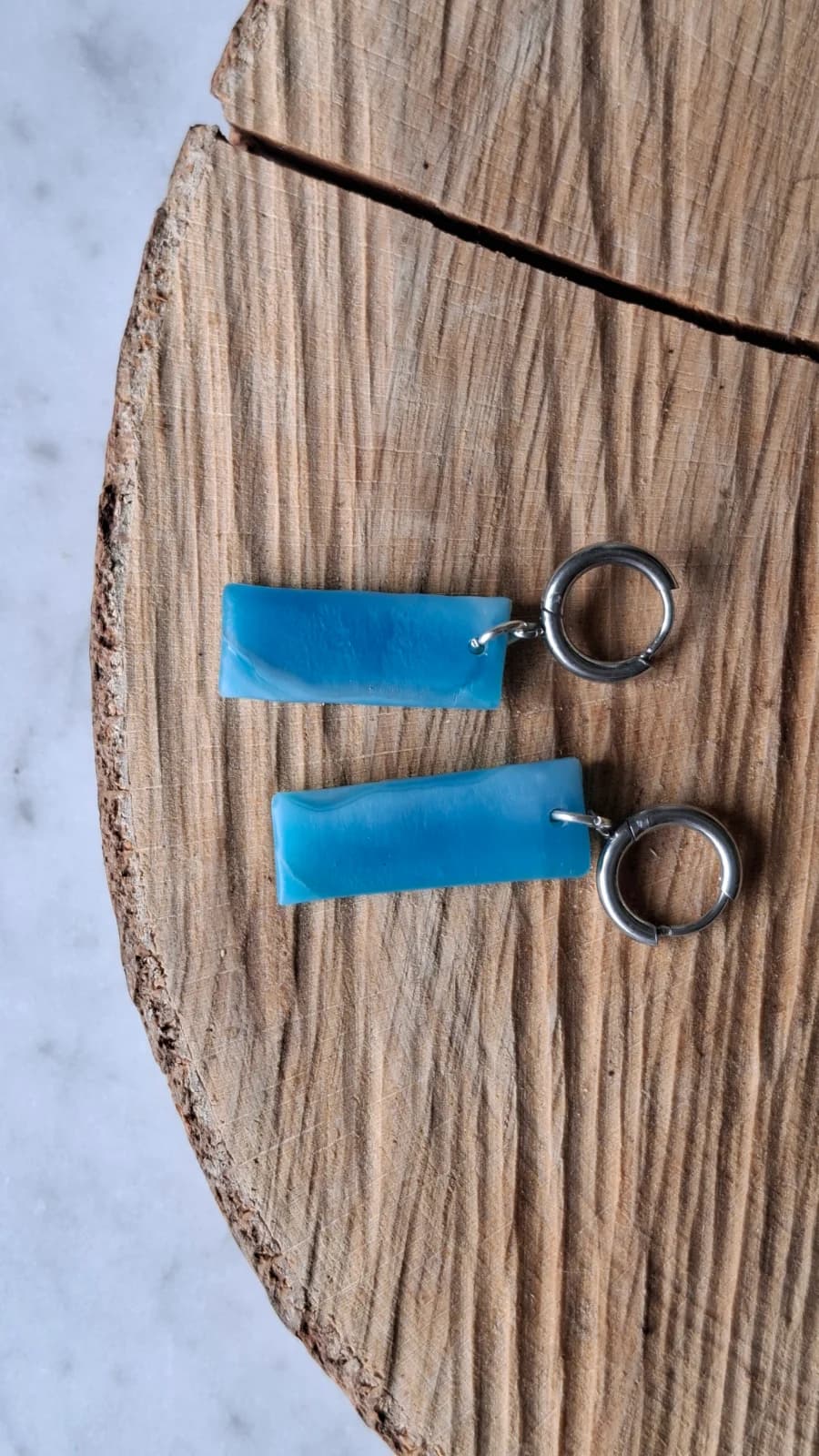 Paire de boucles d'oreilles dormeuses en verre recyclé bleu posées à plat sur une tranche de bois.