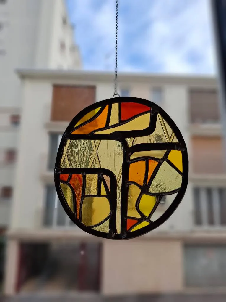 Médaillon en vitrail rond abstrait aux tons jaunes et orangés suspendu par une fine chaîne métallique.