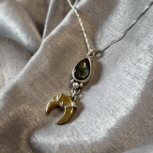 Gros plan sur la goutte de labradorite et la lune dorée du pendentif posé sur tissu argenté
