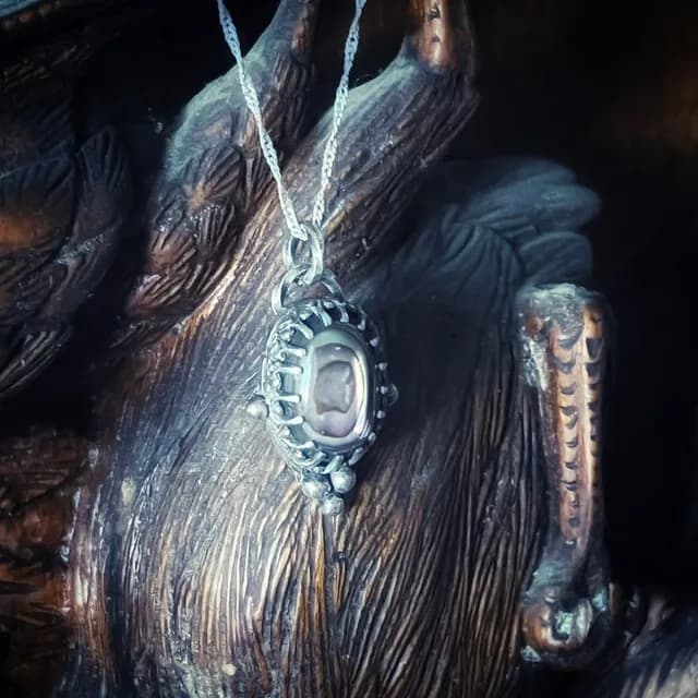Pendentif avec cabochon d'abalone sur chaîne argentée, suspendu devant un support sculpté texturé.