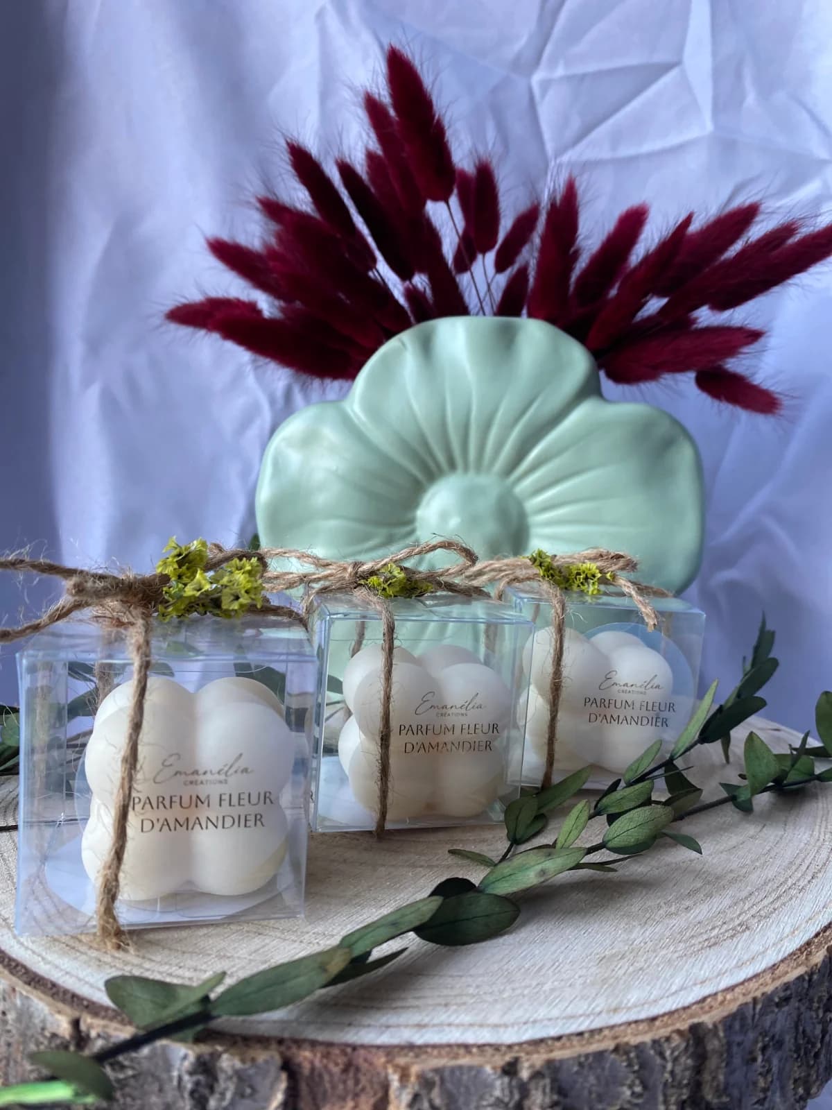 Boîtes transparentes de fondants parfumés blancs avec ficelle et fleurs séchées, disposées sur un rondin de bois.